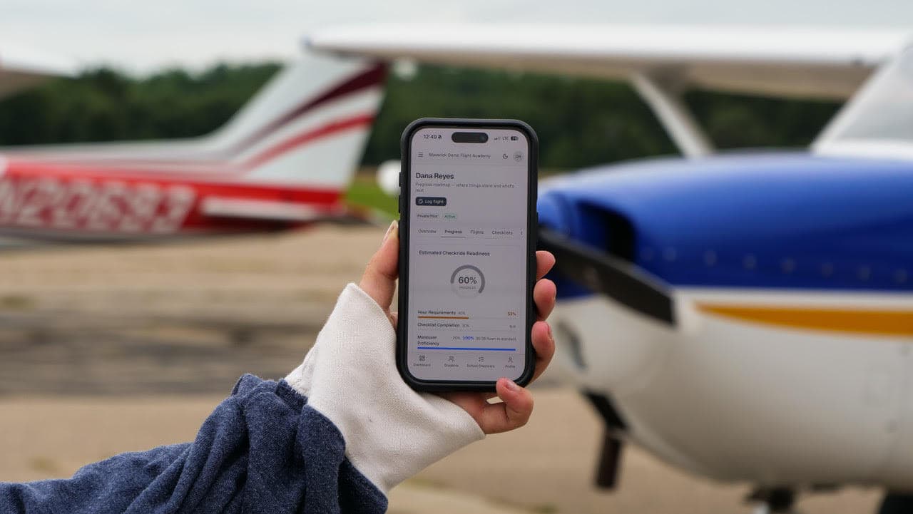A hand holding a phone on an airport ramp between two training airplanes. The phone shows a student's estimated checkride readiness in Maverick.